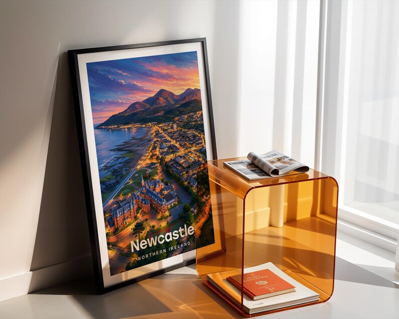 Newcastle cityscape poster presenting the sandy beach, the peak of Slieve Donard, and the Mourne Mountains above the seaside town in bold illustrative colour with clean white NEWCASTLE typography overhead. An open sky of drifting clouds and circling birds fills the upper composition. Framed in black, leaned in a white-walled corner beside an amber acrylic side table and an open magazine in warm shadow light.