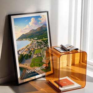 Newcastle cityscape poster presenting Slieve Donard rising above the promenade and sandy beach, the Mourne Mountains rolling to the horizon, and the seaside town below in bold illustrative colour with clean white NEWCASTLE typography overhead. An open sky of drifting clouds and circling birds fills the upper composition. Framed in black, leaned in a white-walled corner beside an amber acrylic side table and an open magazine in warm shadow light.