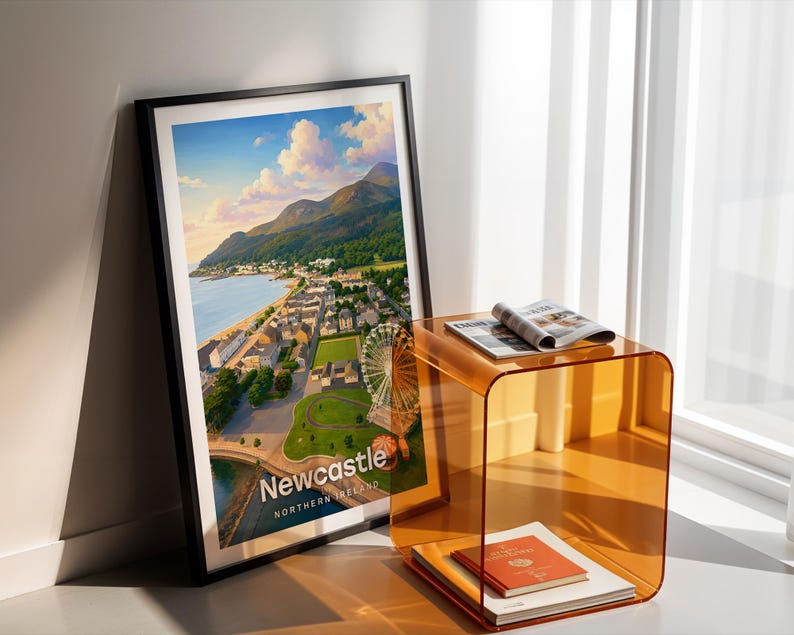 Newcastle cityscape poster presenting Slieve Donard rising above the promenade and sandy beach, the Mourne Mountains rolling to the horizon, and the seaside town below in bold illustrative colour with clean white NEWCASTLE typography overhead. An open sky of drifting clouds and circling birds fills the upper composition. Framed in black, leaned in a white-walled corner beside an amber acrylic side table and an open magazine in warm shadow light.