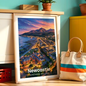 Newcastle poster capturing the sweeping sandy beach, the dramatic peak of Slieve Donard rising from the shoreline, and the Mourne Mountains stretching across the horizon above the seaside town in warm illustrative colour. Bold white NEWCASTLE typography anchors the composition beneath drifting clouds. Framed in light oak, leaned against a teal wall beside a white cabinet and terracotta planted pots.