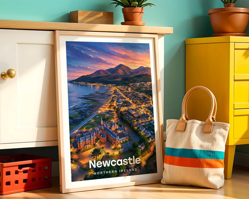 Newcastle poster capturing the sweeping sandy beach, the dramatic peak of Slieve Donard rising from the shoreline, and the Mourne Mountains stretching across the horizon above the seaside town in warm illustrative colour. Bold white NEWCASTLE typography anchors the composition beneath drifting clouds. Framed in light oak, leaned against a teal wall beside a white cabinet and terracotta planted pots.