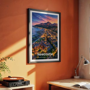 Modern Newcastle city art bringing the sweeping beach promenade, the dramatic Slieve Donard peak, and the Mourne Mountains rolling above the seaside town to life in warm illustrated tones with bold white NEWCASTLE typography. Framed in black and hung on a terracotta textured wall beside a vintage record player, yellow lamp, and open book in a characterful reading corner.