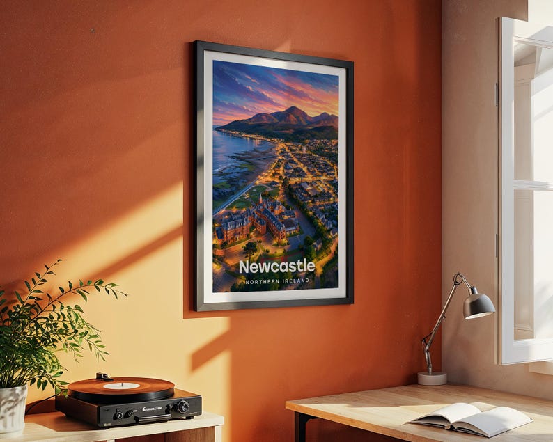 Modern Newcastle city art bringing the sweeping beach promenade, the dramatic Slieve Donard peak, and the Mourne Mountains rolling above the seaside town to life in warm illustrated tones with bold white NEWCASTLE typography. Framed in black and hung on a terracotta textured wall beside a vintage record player, yellow lamp, and open book in a characterful reading corner.