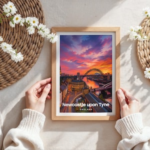 Capturing Newcastle upon Tyne in warm illustrative colour, this framed print showcases the Tyne Bridge spanning the River Tyne with Grey Street's sweeping Georgian facades and Grey's Monument rising behind, finished with bold white NEWCASTLE UPON TYNE typography and birds drifting through an open illustrated sky. Oak frame laid flat on a cream linen surface with a rattan oval placemat, white daisy blooms, and hands holding the frame in soft natural daylight.