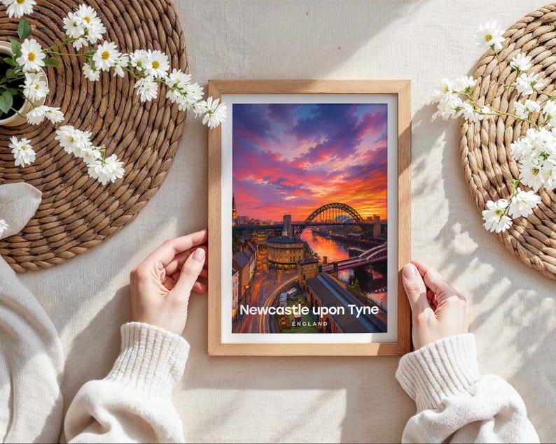Capturing Newcastle upon Tyne in warm illustrative colour, this framed print showcases the Tyne Bridge spanning the River Tyne with Grey Street's sweeping Georgian facades and Grey's Monument rising behind, finished with bold white NEWCASTLE UPON TYNE typography and birds drifting through an open illustrated sky. Oak frame laid flat on a cream linen surface with a rattan oval placemat, white daisy blooms, and hands holding the frame in soft natural daylight.