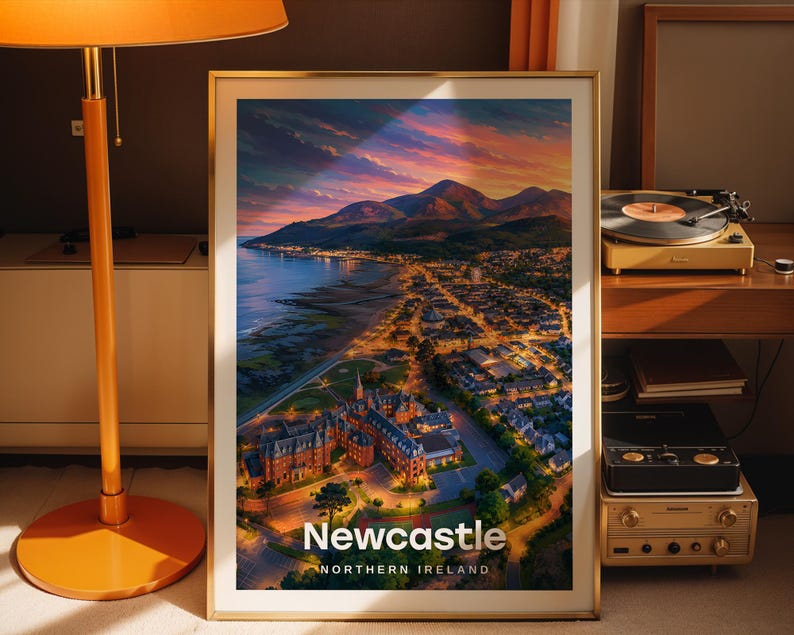 Designed as a Newcastle art print, this high-detail coastal cityscape illustration highlights the sandy beach, the towering peak of Slieve Donard, and the Mourne Mountains horizon under an open illustrated sky. Bold white NEWCASTLE typography anchors the scene. Set in a gold frame leaned against a dark taupe wall, a warm-shaded floor lamp and vintage record player completing the vignette.