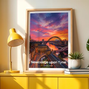 Framed Newcastle upon Tyne wall art print featuring the iconic Tyne Bridge arching over the River Tyne with the curved Georgian terraces of Grey Street and Grey's Monument lantern rising on the hill behind. High-detail illustrated cityscape with rich warm colour, bold white NEWCASTLE UPON TYNE typography, and an open illustrated sky with drifting clouds. Oak frame leaning against a cream wall beside a yellow console, stacked books, and a green plant in a white pot.