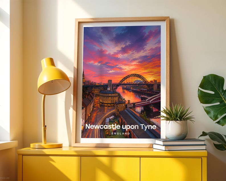 Framed Newcastle upon Tyne wall art print featuring the iconic Tyne Bridge arching over the River Tyne with the curved Georgian terraces of Grey Street and Grey's Monument lantern rising on the hill behind. High-detail illustrated cityscape with rich warm colour, bold white NEWCASTLE UPON TYNE typography, and an open illustrated sky with drifting clouds. Oak frame leaning against a cream wall beside a yellow console, stacked books, and a green plant in a white pot.