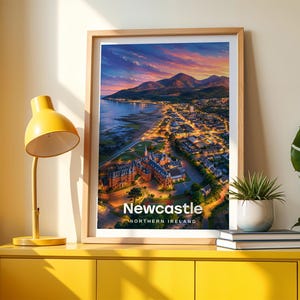 Framed Newcastle wall art print featuring a high-detail illustrated coastal scene with the sandy beach and promenade, Slieve Donard rising steeply from the shore, and the Mourne Mountains rolling along the horizon. Bold white NEWCASTLE typography stands out against an open illustrated sky with drifting clouds and circling birds. Displayed in a light oak frame on a cream wall beside a yellow console and warm lamp light.