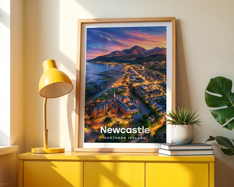 Framed Newcastle wall art print featuring a high-detail illustrated coastal scene with the sandy beach and promenade, Slieve Donard rising steeply from the shore, and the Mourne Mountains rolling along the horizon. Bold white NEWCASTLE typography stands out against an open illustrated sky with drifting clouds and circling birds. Displayed in a light oak frame on a cream wall beside a yellow console and warm lamp light.