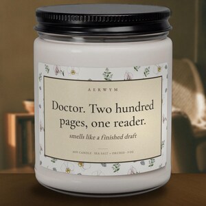 May include: A 9 oz soy candle in a clear glass jar with a black lid. The label features a cream background with a floral border and the text: AERWYM, Doctor. Two hundred pages, one reader. smells like a finished draft, SOY CANDLE SEA SALT + ORCHID 9 OZ.