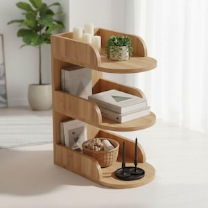 May include: A light-colored, three-tiered wooden bookshelf with rounded shelves. The top shelf holds white candles and a small potted plant. Books, a woven basket, and candles are on the other shelves. The bookshelf is in a well-lit room.