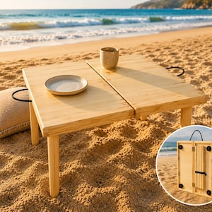 Puede incluir: Una mesa de picnic plegable de madera clara con un plato y una taza encima, colocada en una playa de arena. Una almohada beige con un asa negra está al lado de la mesa. La mesa se pliega para facilitar el almacenamiento y el transporte.