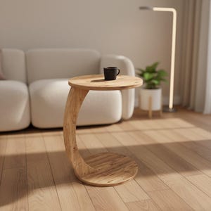 May include: A C-shaped side table crafted from light-toned wood with a visible grain pattern, featuring a circular top and a matching circular base connected by a curved support pillar. A small black mug sits on the surface of the table.