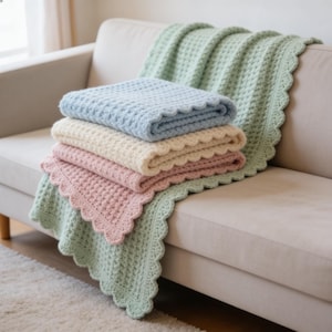 Puede incluir: Una pila de cuatro mantas tejidas a ganchillo con un patrón de punto de gofre texturizado y bordes festoneados. Las mantas son de color verde salvia claro, azul bebé suave, crema y rosa empolvado, cada una doblada cuidadosamente para mostrar el tejido.