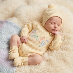 May include: A pale yellow knitted baby romper featuring long sleeves, a ribbed texture, and three buttons at the bottom. The chest displays the name Emily embroidered in pastel purple, green, and blue script. Includes a matching beanie and booties.