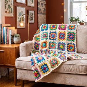 May include: A crochet blanket featuring a repeating granny square pattern with vibrant multi-colored centers in shades of blue, green, yellow, pink, and red, all framed by a cream-colored yarn border, draped over a light-colored upholstered sofa.