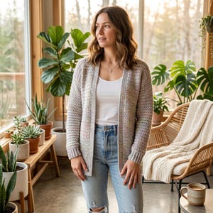 May include: A light-colored, open-front cardigan with a textured knit pattern. The cardigan is worn over a white top and paired with blue jeans. The background features a sunroom with plants and a wicker chair.