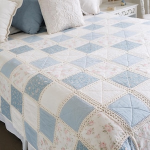May include: A patchwork quilt featuring a repeating pattern of solid light blue squares, white squares, and squares with a pink floral print. Each square is joined by a cream-coloured crocheted border, and the quilt is draped over a bed.