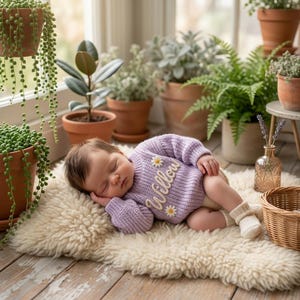 May include: A lavender long-sleeve knit sweater featuring the name Willow embroidered in white cursive script, accented with two small white daisy flowers. The sweater is worn by a sleeping infant resting on a cream-colored sheepskin rug.