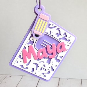 May include: A purple and white hanging ornament shaped like a notebook with a marbled pattern. The name Maya is written in raised pink letters across the center. A small, light pink and yellow pencil charm is attached to the top of the notebook.