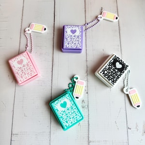 May include: Three rectangular silicone cases with speckled patterns in pink, purple, and teal, each featuring a central heart shape. Each case includes a matching ball chain attached to a small, yellow and white pencil-shaped charm.