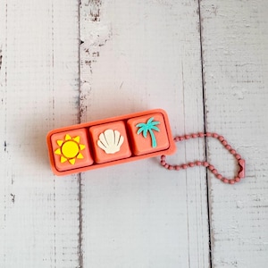 May include: A rectangular coral-colored plastic keycap holder featuring three square slots. Each slot contains a raised icon: a yellow sun, a white seashell, and a teal palm tree. A matching coral-colored ball chain is attached to the side.