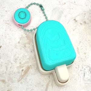 May include: A turquoise popsicle-shaped keychain with a white holder and stick. A pink kiwi-shaped charm with a green beaded chain is attached. The keychain is on a light-colored, textured surface.