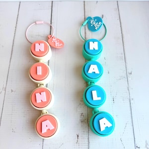 May include: Two vertical pacifier clips featuring circular silicone beads. The left clip is cream with coral beads spelling NINA, and the right is teal with matching beads spelling NALA. Each has a circular tag with the text MNM attached to a ring.