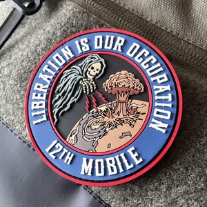 May include: A circular morale patch with a red border and a blue ring featuring the white text "LIBERATION IS OUR OCCUPATION" and "12TH MOBILE". The centre depicts a skeleton figure emitting red lightning bolts toward a planet with a mushroom cloud.