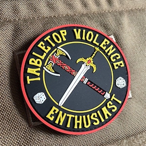 May include: A circular morale patch with a black background and a red border. It features a crossed axe and sword, a six-sided die, and a twenty-sided die. The yellow text reads TABLETOP VIOLENCE ENTHUSIAST, arranged in a circular pattern.