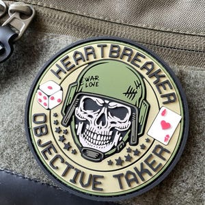 May include: A circular morale patch featuring a skull wearing an olive green helmet with the text WAR LOVE. The patch includes a white die, a playing card with a red heart, and the text HEARTBREAKER OBJECTIVE TAKER in black lettering.