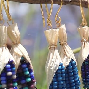 May include: A collection of dangling earrings shaped like miniature corn cobs. Each earring features a cluster of small, multi-colored glass beads in shades of blue, purple, and white, topped with light beige husks secured by thin gold-toned wire.