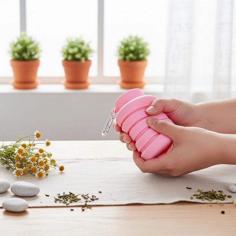 Puede incluir: Una botella de agua plegable de silicona rosa con un dise&ntilde;o acanalado tipo acorde&oacute;n y un mosquet&oacute;n de metal plateado sujeto al cuello, lo que permite un almacenamiento compacto y un transporte sencillo durante viajes o actividades al aire libre.