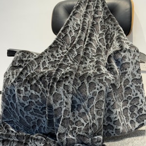 May include: A plush, faux fur throw blanket featuring a dark gray and black animal print pattern. The fabric has a thick, textured pile with a soft appearance, draped over a black chair. The pattern consists of irregular, interconnected shapes.