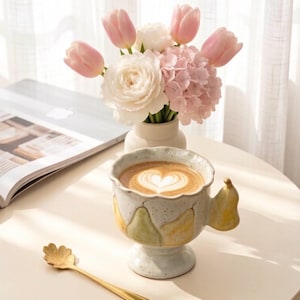Puede incluir: Una taza de cerámica con borde festoneado y asa en forma de pera, con acabado moteado y motivos de peras pintados, llena de café con arte latte en forma de corazón, acompañada de una cuchara dorada con mango en forma de hoja.