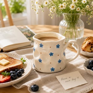 Puede incluir: Una taza de cerámica color crema con borde irregular y ondulado, que incluye un asa y un patrón repetitivo de pequeñas flores azules y puntos marrones. La taza contiene un líquido marrón claro y se apoya sobre una superficie de tela blanca.