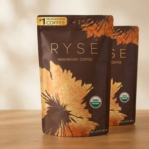 May include: Two brown pouches of RYSE Mushroom Coffee. The front pouch has a large gold floral design and the text "RYSE MUSHROOM COFFEE". The pouches are labeled "USDA ORGANIC" and "NET WT 6.35 OZ (180 G)".