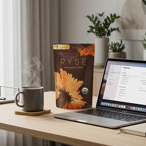 May include: A home office setup with a laptop, coffee, and a bag of RYSE mushroom coffee. A steaming mug sits on a wooden coaster. A notebook, pen, and headphones are also on the desk. The laptop screen displays a productivity app.