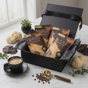 May include: A black gift box with four packets of RYSE mushroom coffee, a cup of coffee, coffee beans, and a gold scoop. The packets are brown with a sunflower design. Other items include a small succulent, and various mushrooms.