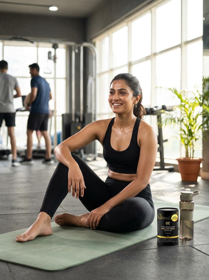 May include: A woman in black athletic wear sits on a green yoga mat in a gym. She wears a black sports bra and leggings. A black and gold container and a water bottle are next to her. The text on the container reads "BE PURE HIMALAYAN SILANT GOLD CRANBERRIES."