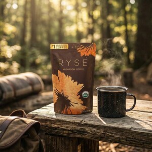 May include: A brown bag of Ryse Mushroom Coffee with a sunflower design and a steaming black speckled enamel mug on a wooden table. A rolled-up blanket, backpack, and tent are in the background, suggesting a camping theme.