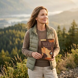May include: A woman stands outdoors, holding a package of Ryse Mushroom Coffee. She wears a green vest, brown sweater, and light-colored pants. The package features a sunflower design. The background includes a forest and mountains.