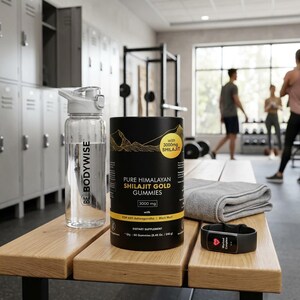 May include: A clear water bottle with a white lid and the text "BE BODYWISE" sits next to a black container of "PURE HIMALAYAN SHILAJIT GOLD GUMMIES". A fitness tracker and a folded gray towel are also on the wooden bench. The background shows a gym setting.