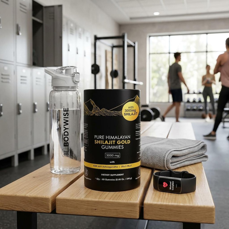 May include: A clear water bottle with a white lid and the text "BE BODYWISE" sits next to a black container of "PURE HIMALAYAN SHILAJIT GOLD GUMMIES". A fitness tracker and a folded gray towel are also on the wooden bench. The background shows a gym setting.