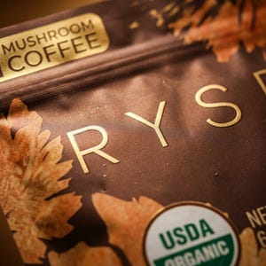 May include: Close-up of a brown package of mushroom coffee. The package has gold lettering that spells out "RYSE" and "Mushroom Coffee". The package also has a USDA Organic seal and states the net weight is 6.35 oz.