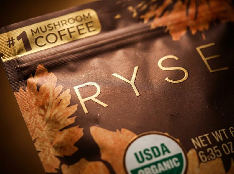 May include: Close-up of a brown package of mushroom coffee. The package has gold lettering that spells out "RYSE" and "Mushroom Coffee". The package also has a USDA Organic seal and states the net weight is 6.35 oz.