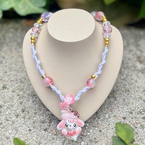 May include: A necklace featuring a white knotted cord, pink and clear faceted beads, and small gold-tone spacer beads. The centerpiece is a pink and white rabbit-shaped charm with a small pink flower accent attached to the top of the charm.