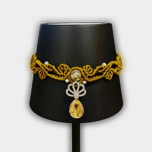 May include: A mustard yellow macrame choker necklace featuring intricate knotted patterns, small silver beads, a central round textured stone, and a silver-toned fan-shaped pendant with a dangling, faceted, teardrop-shaped amber-colored crystal.