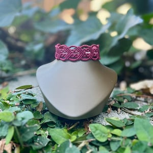 May include: A dark red macrame choker necklace featuring an intricate, repeating heart-shaped knot pattern. The textile jewelry is displayed on a smooth, neutral-toned mannequin bust, showcasing the detailed cord work and consistent knotting.